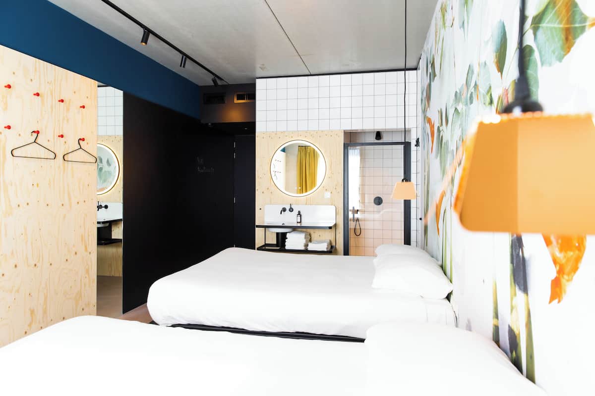 Une chambre d’hôtel moderne avec un grand lit, un mur en bois orné d’un grand miroir rond, une salle de bain avec un lavabo et un espace douche.