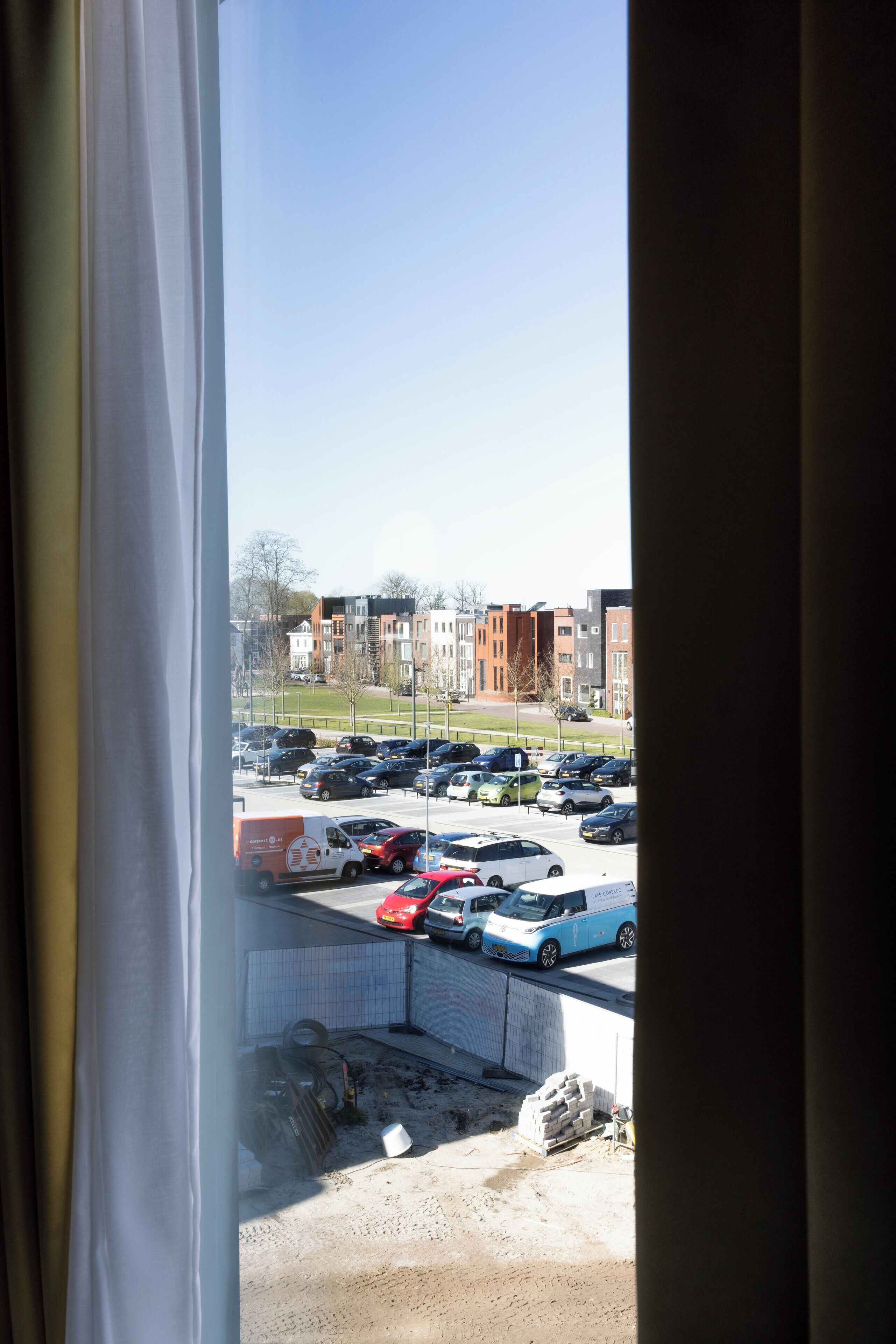 Vista a través de una ventana que muestra un estacionamiento con varios coches, una obra en construcción y edificios residenciales al fondo.