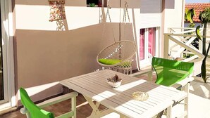 Une terrasse extérieure avec une table et des chaises blanches, une décoration en macramé suspendue, et une vue sur la maison en arrière-plan.