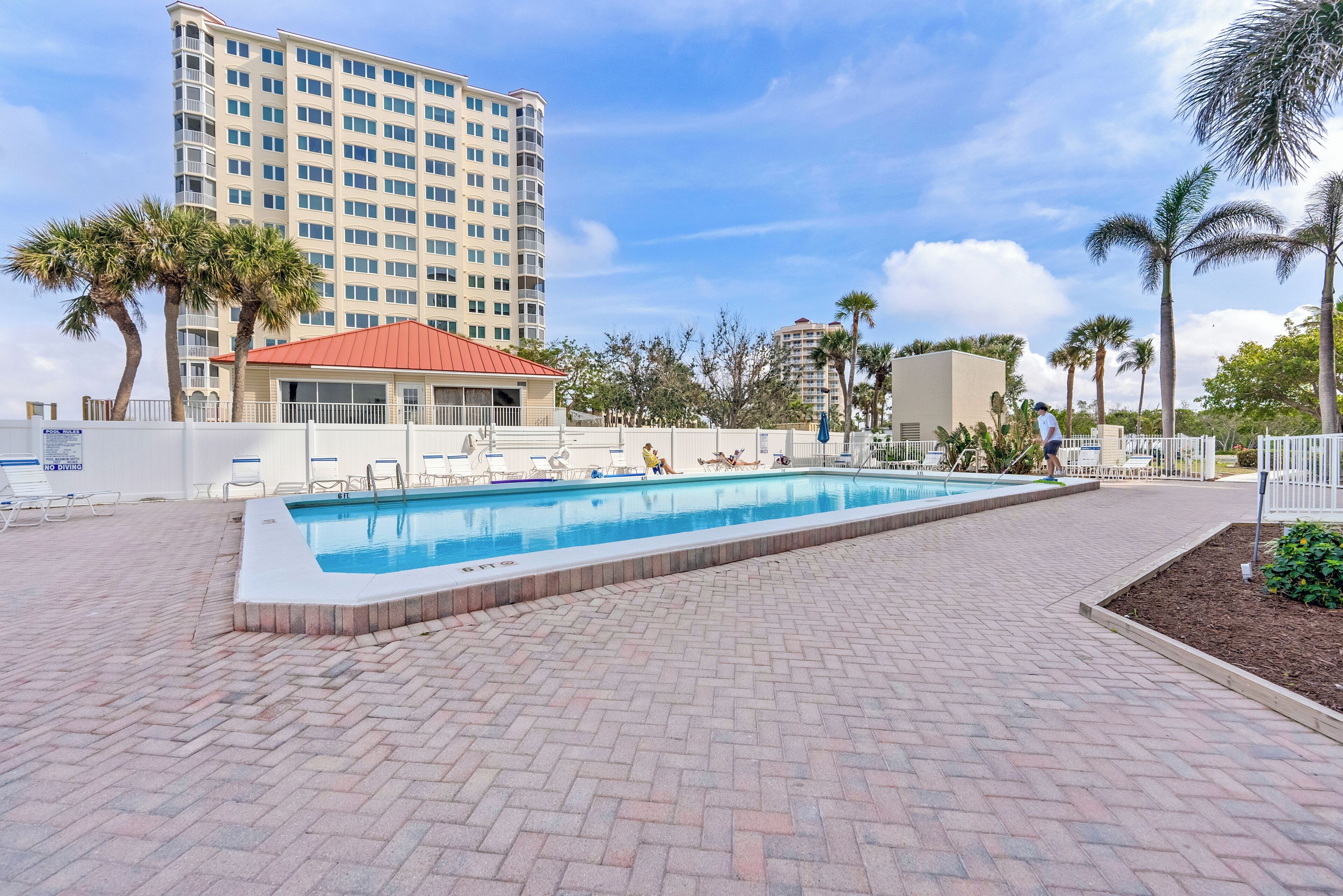 A swimming pool with lounge chairs, a red-roofed building, palm trees, and a tall hotel in the background.