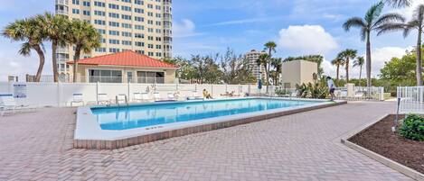 A swimming pool with lounge chairs, a red-roofed building, palm trees, and a tall hotel in the background.
