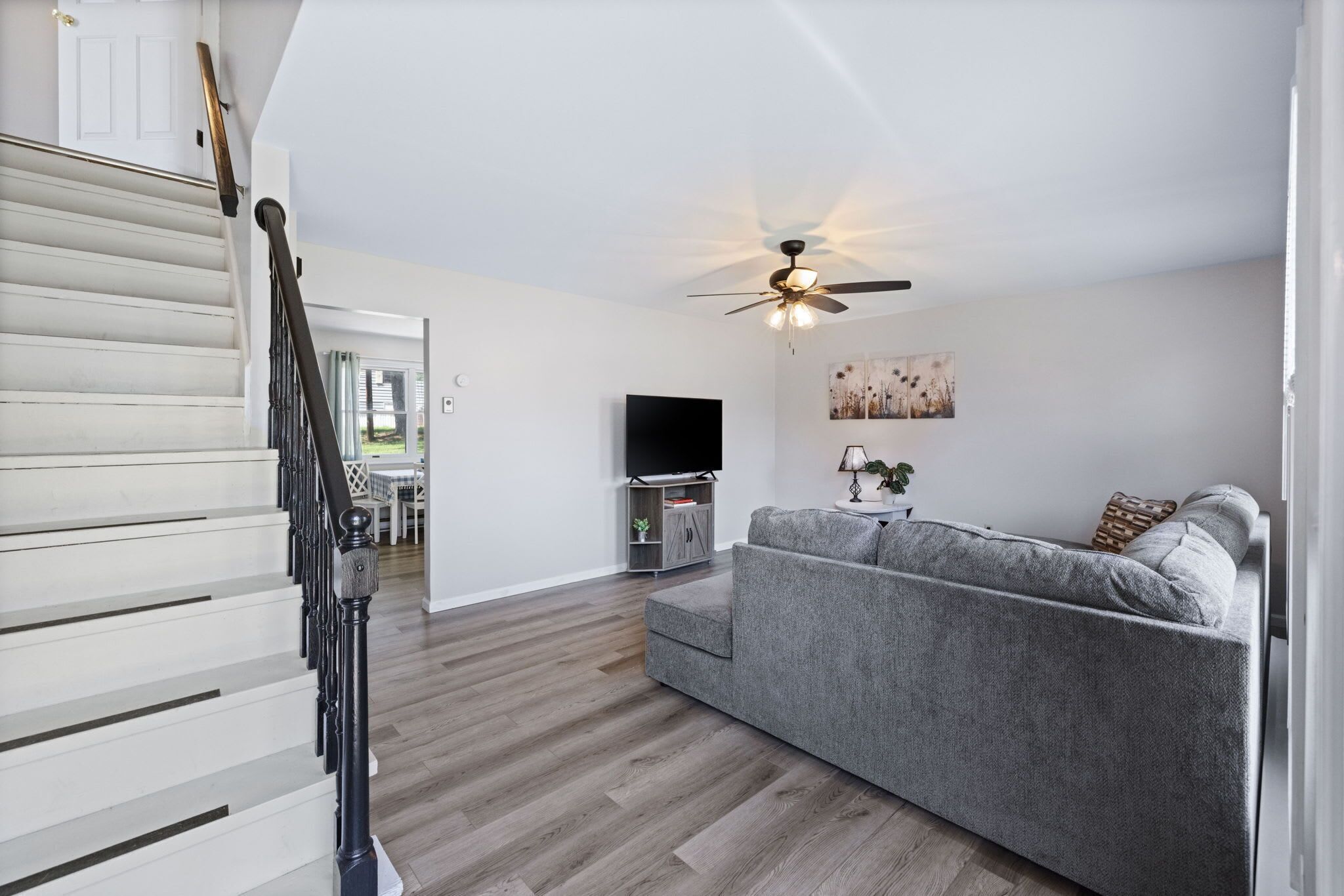 A modern living room with a grey sofa, wooden flooring, a ceiling fan, and a flat-screen TV.
