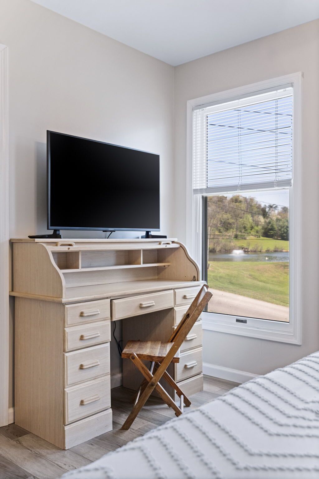 A bedroom with a wooden desk, a television, a chair, and a window with blinds.