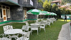 A courtyard with white metal tables and chairs, green and white umbrellas, and a stone building with balconies. - Gagan Resort (Dharamshala)