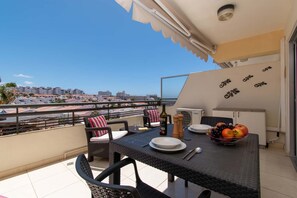 Ein Balkon mit Korbmöbeln, einem Tisch gedeckt mit Tellern, einer Flasche und einer Obstschale.
