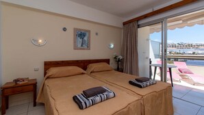 Une chambre d’hôtel avec deux lits, une tête de lit en bois, une table de chevet, une petite table avec un livre, un tableau au mur et un balcon avec des chaises et une table.