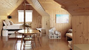 Un dormitorio acogedor en el ático, con techo inclinado de madera, una cama, un escritorio y un pequeño estante blanco.