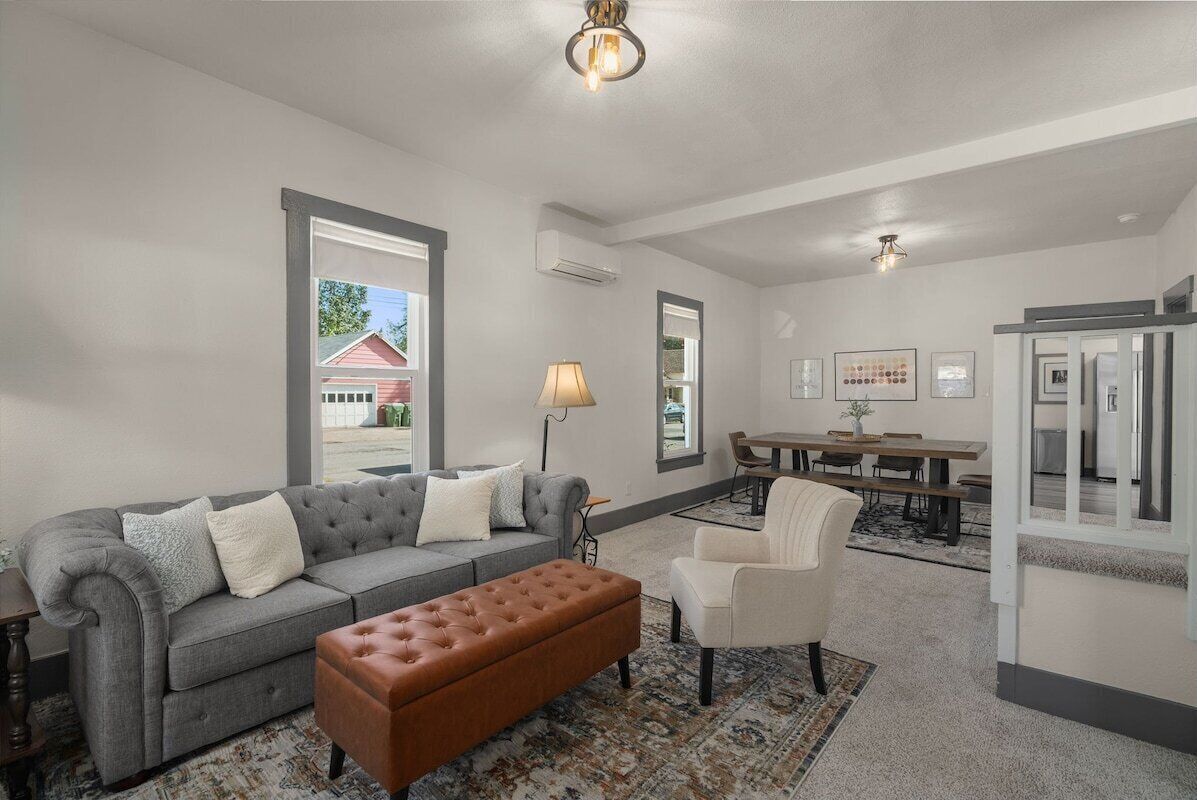 Ein Wohnzimmer mit grauem Sofa, braunem Hocker und einem weißen Sessel. Es gibt eine Essecke mit Tisch und Stühlen.