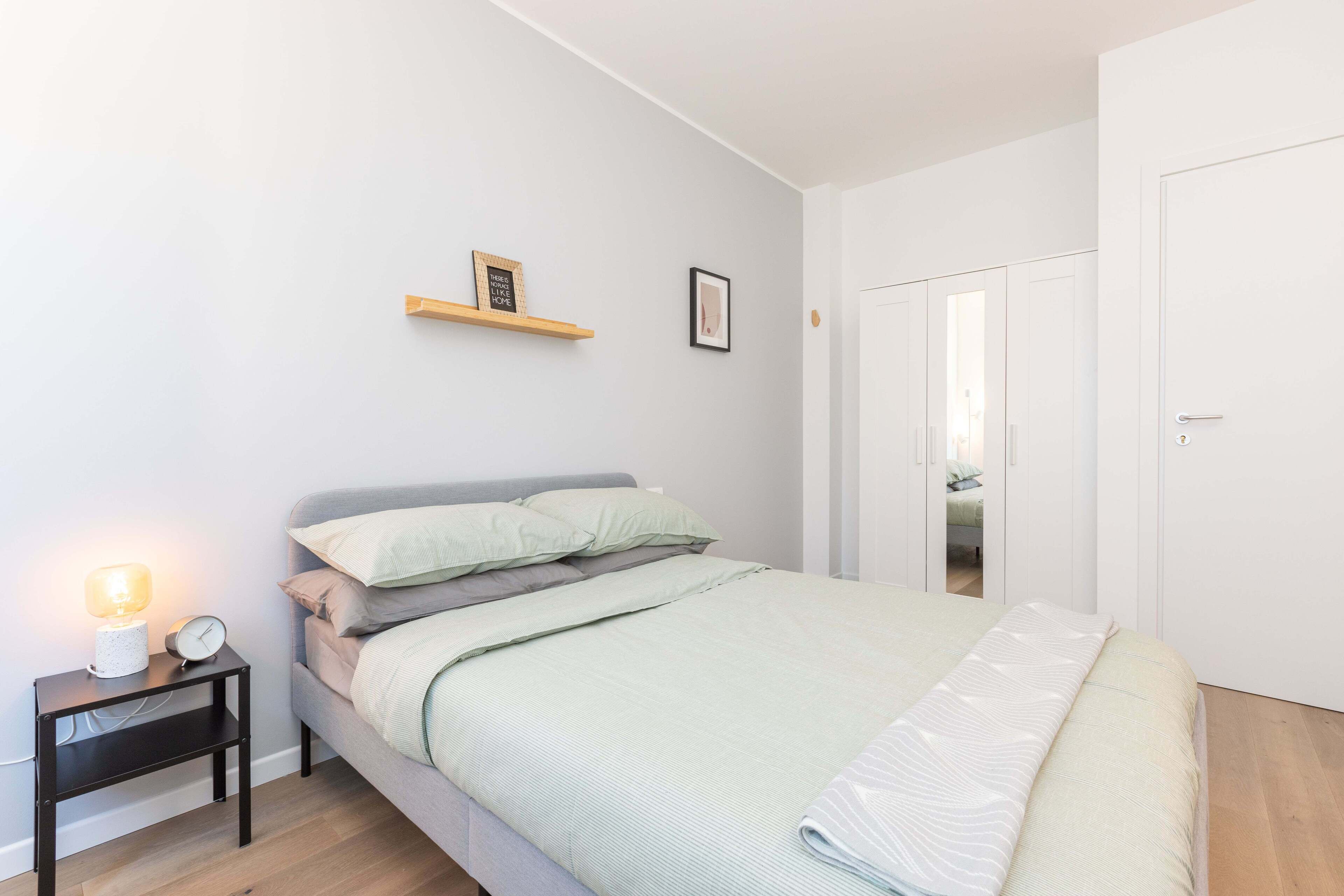 A bedroom with a bed, a nightstand with a lamp and clock, a shelf with a picture, and a door.