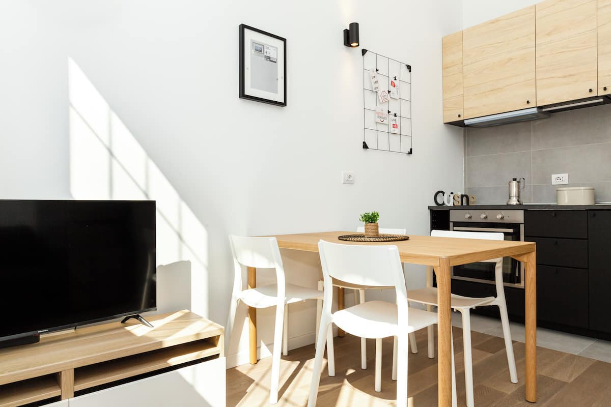 A modern kitchen with wooden cabinets, a dining table with white chairs, a TV, and a wall-mounted light fixture.