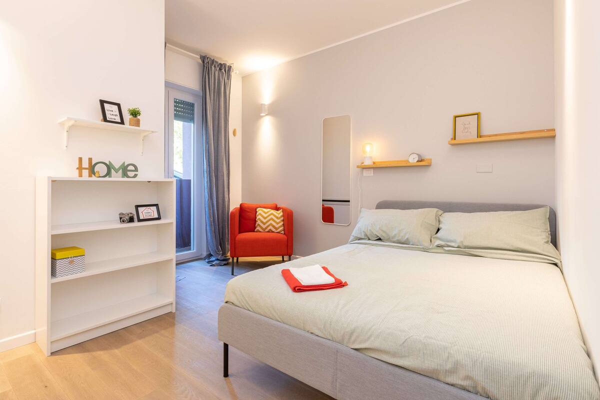 A modern bedroom with a bed, a chair, a shelf with a 'Home' sign, and a mirror.