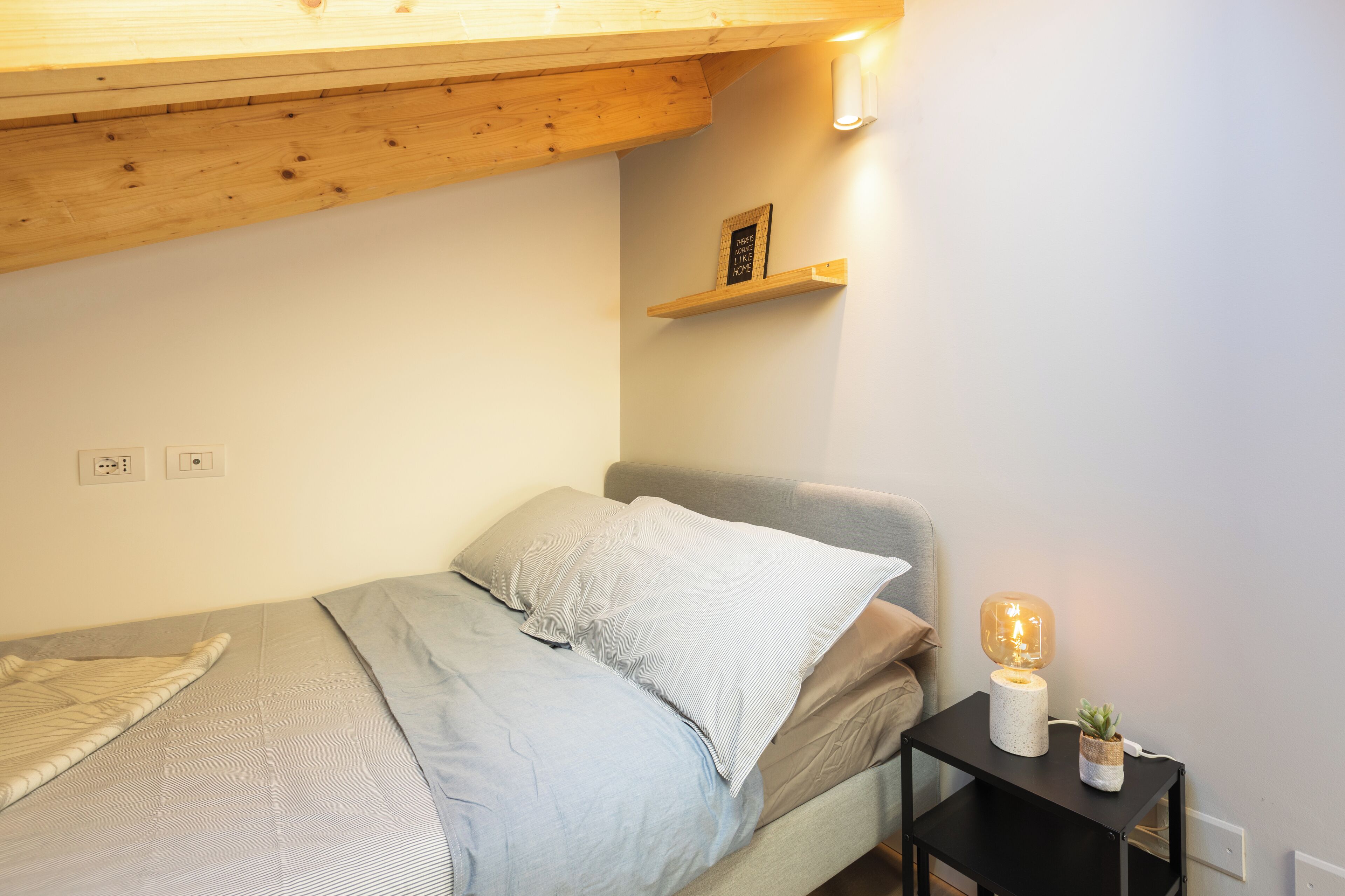 Ein kleines, aufgeräumtes Schlafzimmer mit einem Bett, einem Nachttisch mit Lampe und einem Regal mit einem Bild.