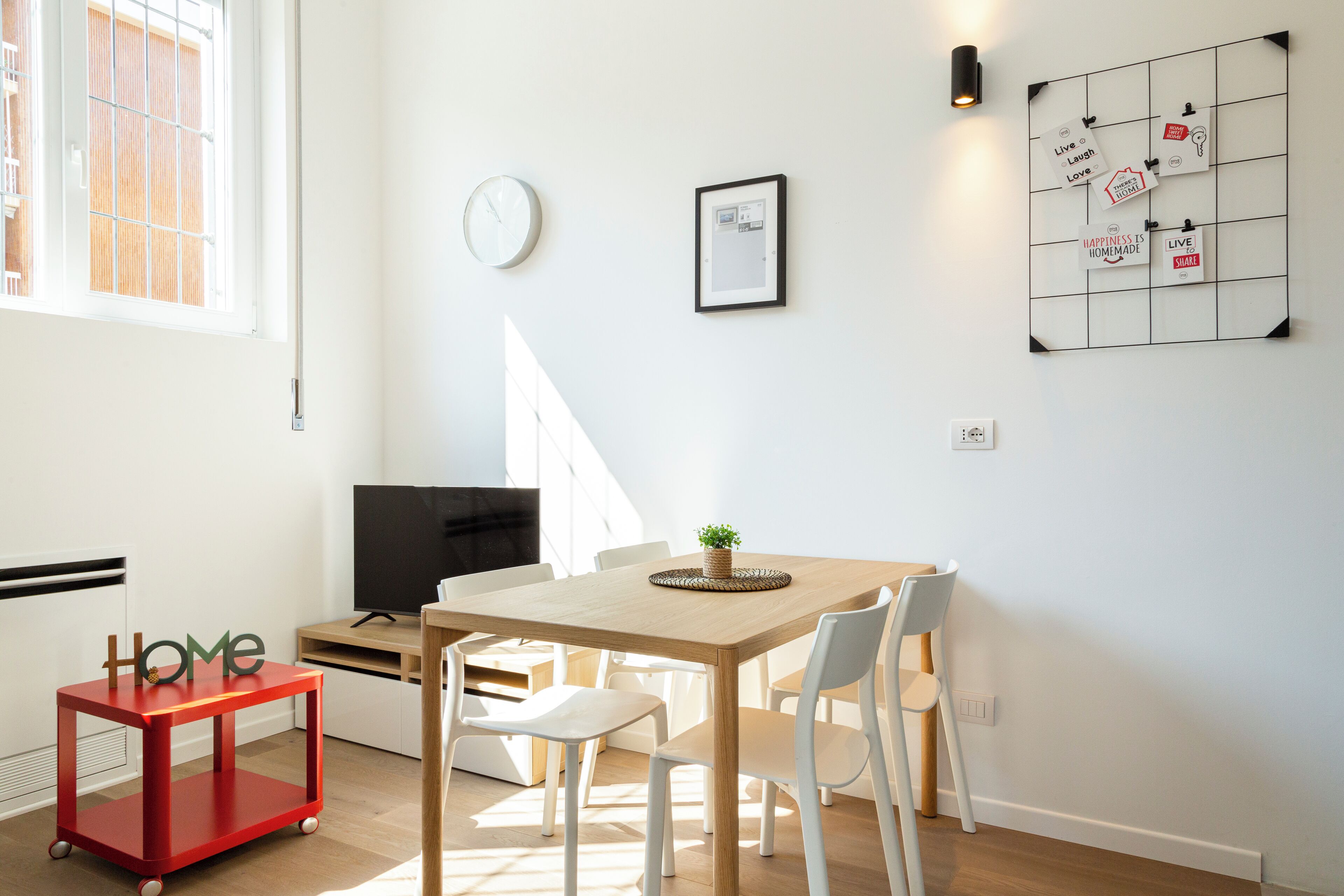 Ein modernes Wohnzimmer mit einem Holzes Esstisch, weißen Stühlen, einem roten Beistelltisch, einem Fernseher und einer Wandleuchte.