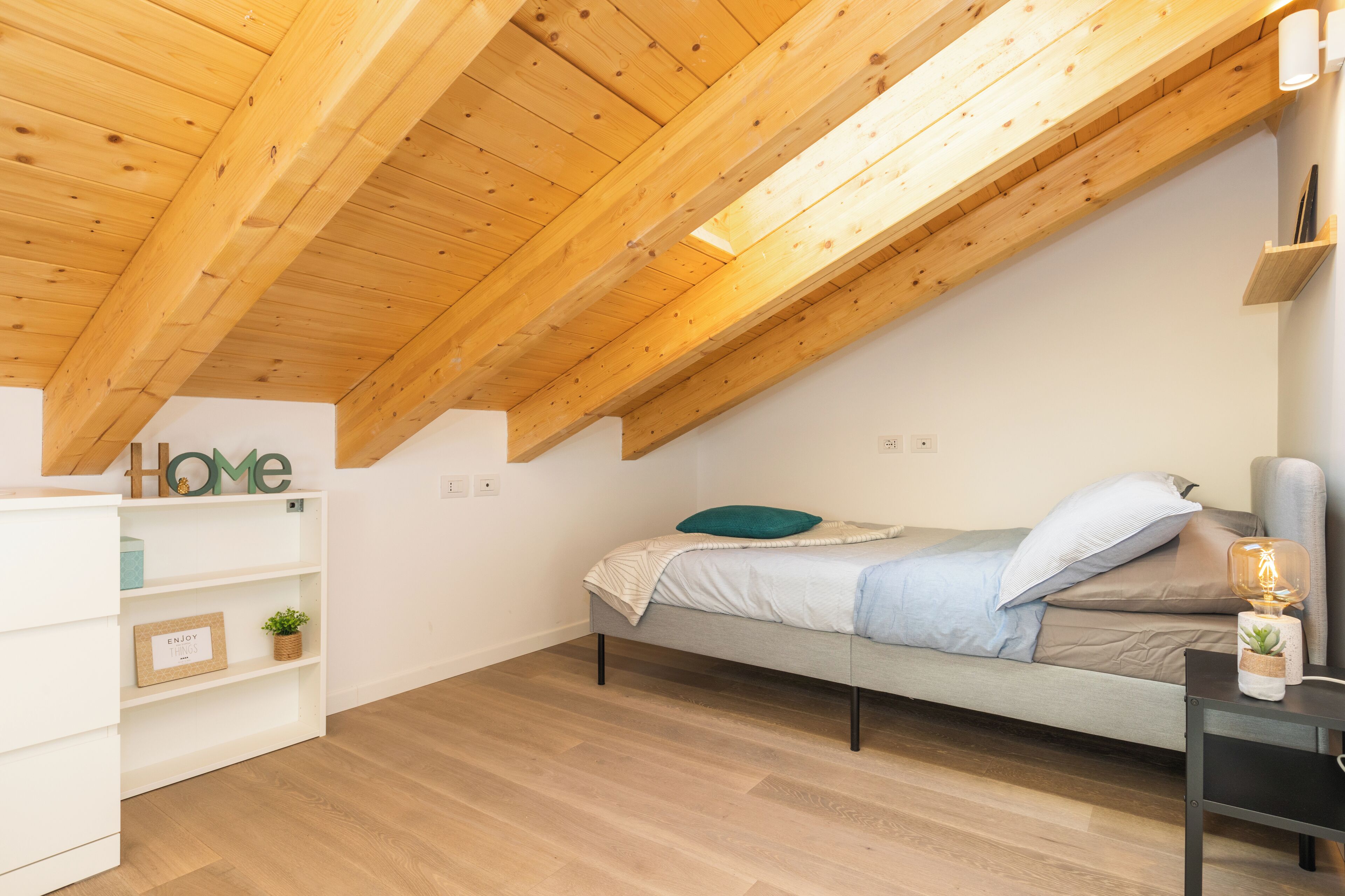 Un dormitorio con techo inclinado, una cama, una mesita de noche, una estantería y un estante fijado a la pared.