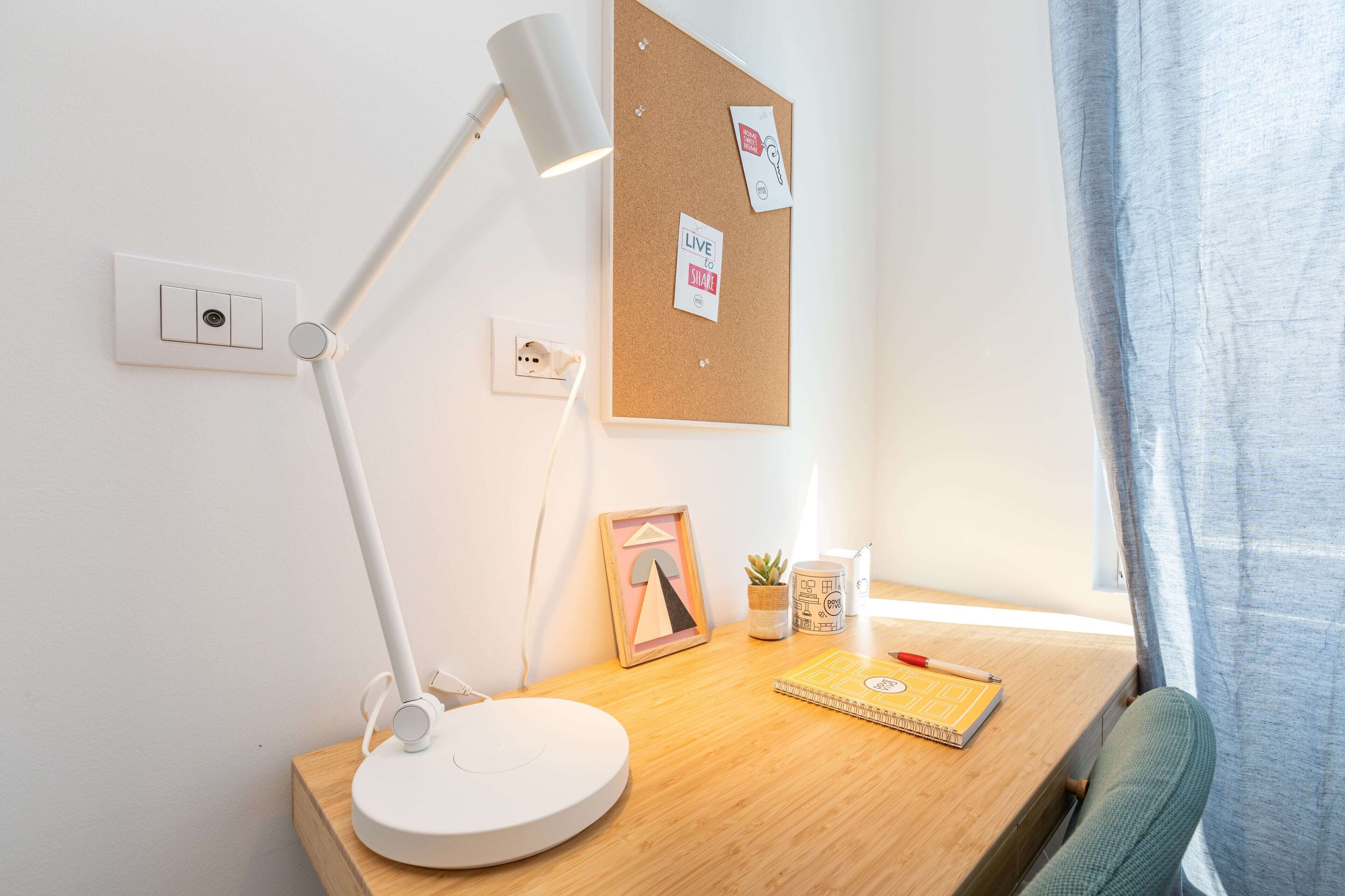 A workspace with a desk, a white desk lamp, a corkboard with notes, a framed picture, a small plant, and a notebook.