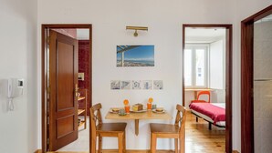Ein Zimmer mit Holzboden, einem Esstisch mit Stühlen, einem Bett mit roter Decke und einem Stadtbild an der Wand.