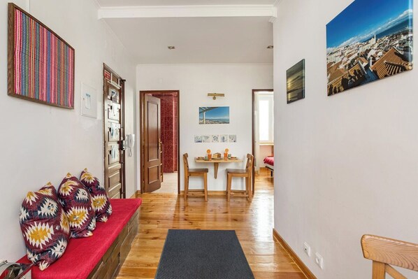 Um corredor com piso de madeira, um sofá vermelho, uma área de refeições com mesa e cadeiras, e obras de arte nas paredes.