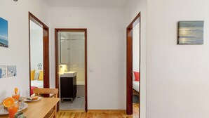 Ein kleines Zimmer mit Holztisch, einem Bett und einem durch eine Glastür einsehbaren Badezimmer.