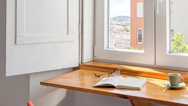 Ein Zimmer mit Holztisch, einem Fenster mit Blick auf ein Gebäude und einer Tasse auf dem Tisch.