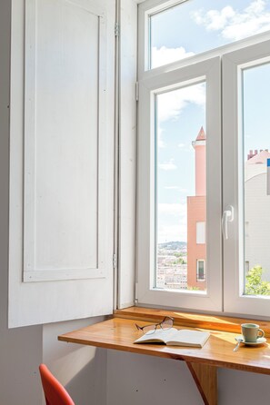 Une pièce avec un bureau en bois, une fenêtre donnant sur un immeuble et une tasse posée sur la table.