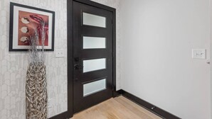 A hallway with a dark wooden door featuring three glass panels, a wall-mounted light switch, and a framed picture with abstract art. - Electric House | Walk to Gulch • Rooftop Hot Tub (Nashville)