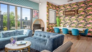 A modern living room with a large window, a dining area with a brick wall, and a vibrant tropical wallpaper. - Electric House | Walk to Gulch • Rooftop Hot Tub (Nashville)