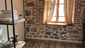 A room with a stone wall, a wooden floor, a metal shelf with towels, and a window with sheer curtains. - Hnameni Stepanavan Guesthouse (Step'anavan)