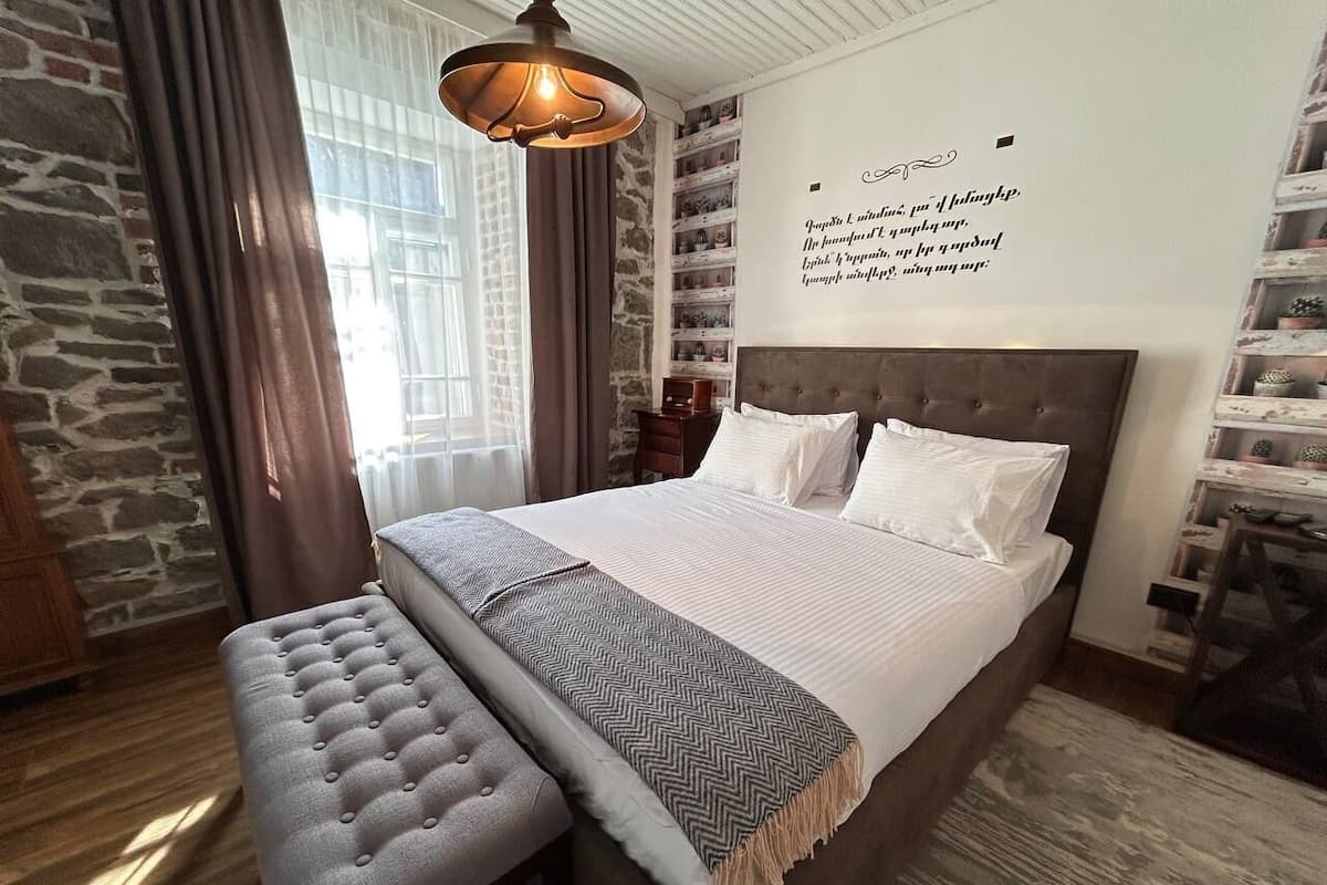Un dormitorio con una cama grande, un banco, un estante en la pared y una ventana con cortinas.