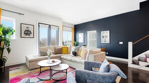 Ein modernes Wohnzimmer mit einer Eckcouch, einem runden Couchtisch und einem blauen Sessel. Große Fenster mit Jalousien, eine Treppe und eine dunkle Akzentwand sind ebenfalls zu sehen.