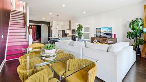 Un salon moderne avec une table basse en verre, des fauteuils de couleur moutarde, un canapé blanc et une cuisine en arrière-plan.