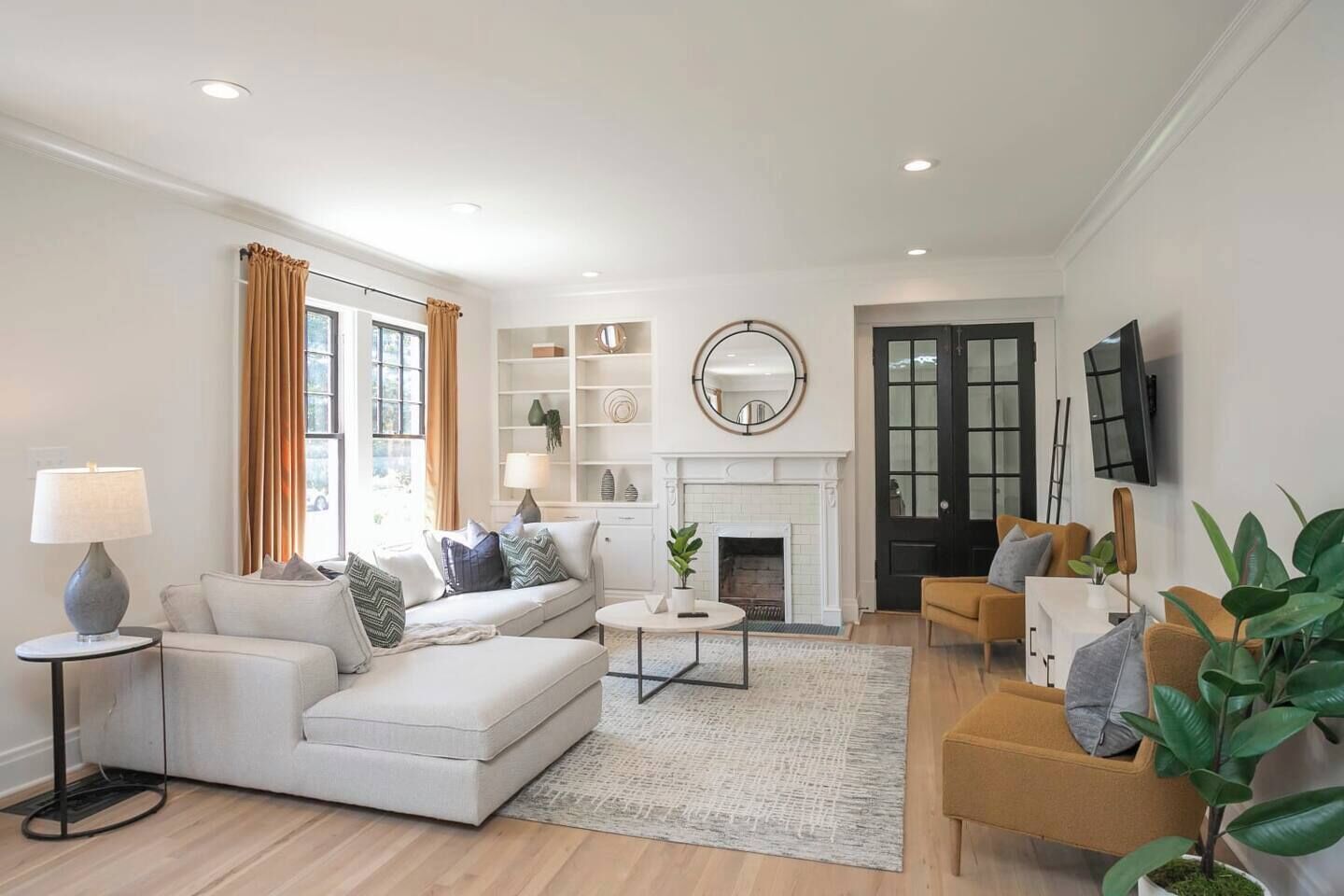 Un salon moderne avec un canapé blanc, une table basse et une cheminée. On y trouve des étagères ornées d’objets décoratifs et un téléviseur fixé au mur.