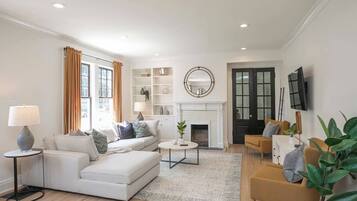 Una sala moderna con un sofá blanco, una mesa de centro y una chimenea. Hay estantes con objetos decorativos y un televisor montado en la pared.