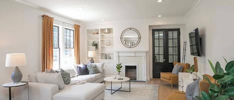 Un salon moderne avec un canapé blanc, une table basse et une cheminée. On y trouve des étagères ornées d’objets décoratifs et un téléviseur fixé au mur.
