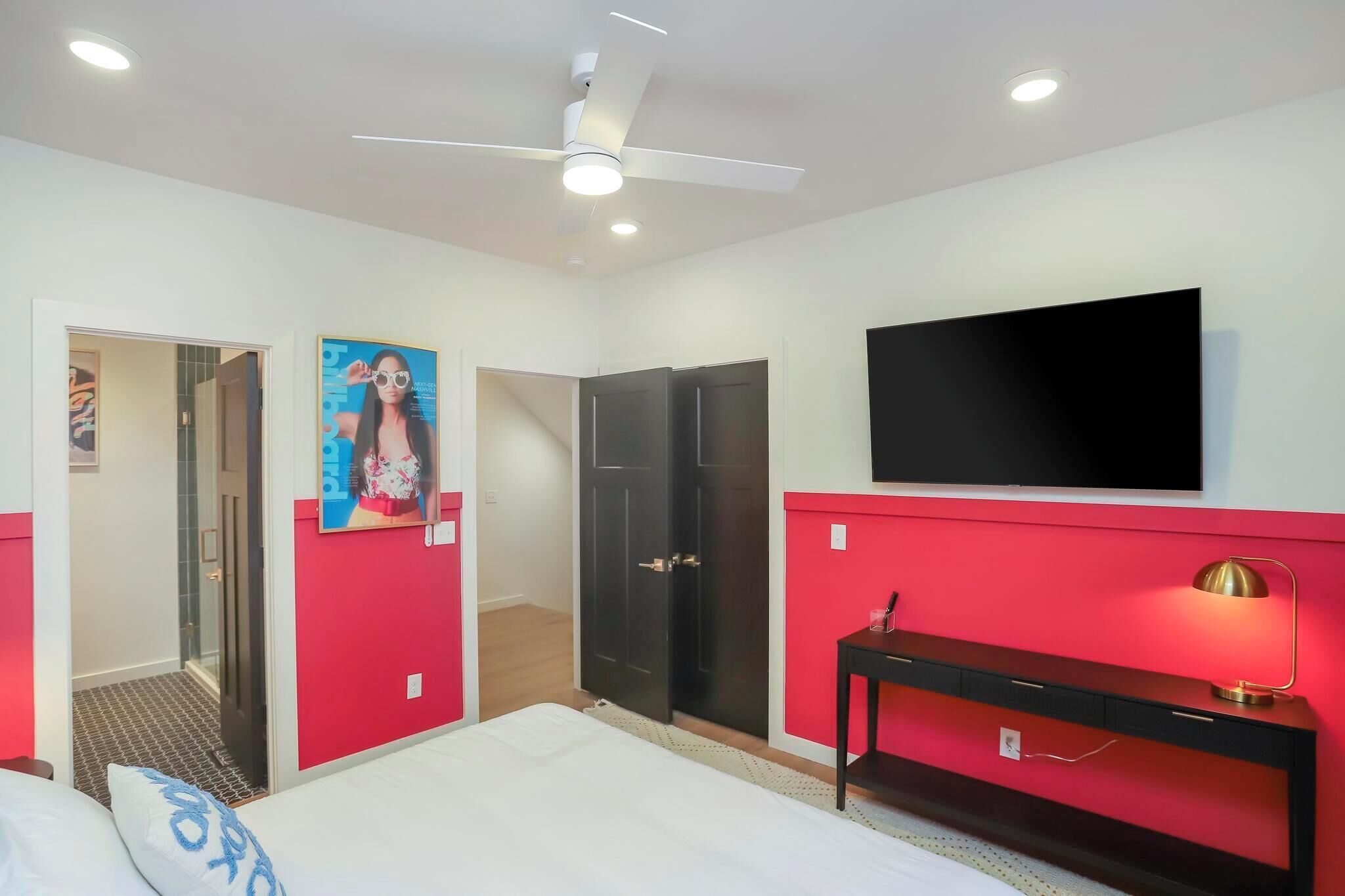 Ein Hotelzimmer mit Bett, Schreibtisch mit Lampe, Wand-Fernseher und Deckenventilator.