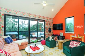 A modern living room with a large sliding glass door leading to an outdoor area, a flat-screen TV mounted on an orange wall, and a variety of seating options including a pink sofa and green armchairs. - Livin' Luxe on Music Row |  Hot Tub & 1mi to DT (Nashville)