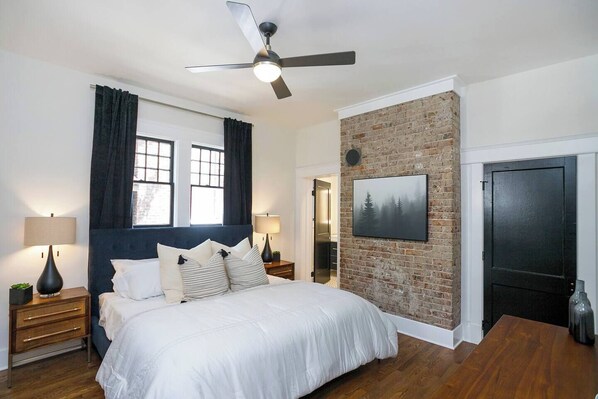 Une chambre avec un grand lit, deux tables de chevet avec des lampes, un ventilateur de plafond, un mur décoratif en briques, une porte et une fenêtre avec des rideaux.