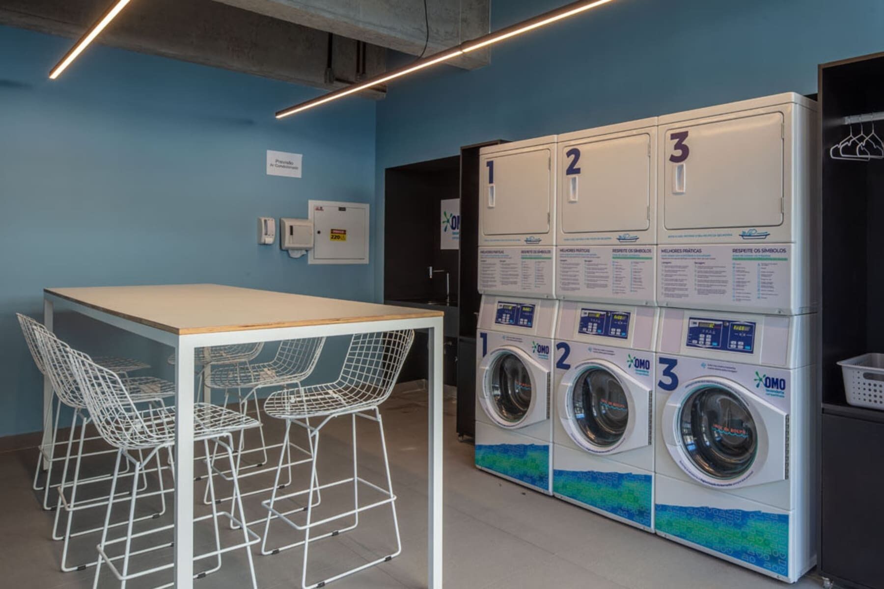 A laundry area with three washing machines, a table with chairs, and a wall-mounted information board.