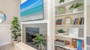 Una sala de estar con chimenea, una estantería llena de libros y juegos de mesa, y un amplio ventanal con vistas al océano.