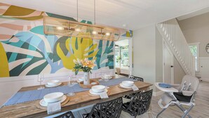 Un comedor con una mesa de madera dispuesta para cuatro personas, un mural abstracto de colores y una lámpara de araña moderna.