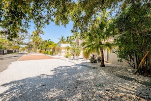 Una calle residencial con camino de grava, palmeras y una casa de fachada color beige.