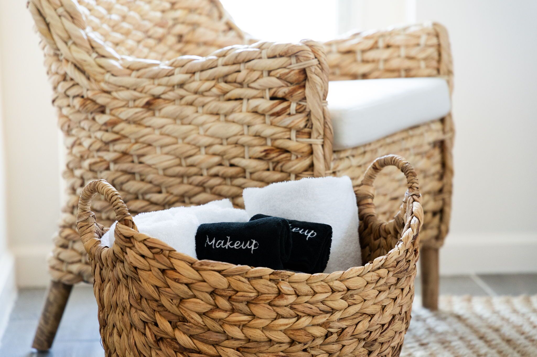 A wicker chair with a white cushion and a basket containing two towels, one labeled 'Makeup'.