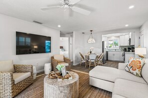 Una sala moderna con televisor de pantalla plana, un comedor con candelabro y una cocina con armarios blancos.