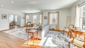 Un salón espacioso con pisos de madera, un sofá gris, sillas de madera, una mesa de centro y ventanas grandes con cortinas.