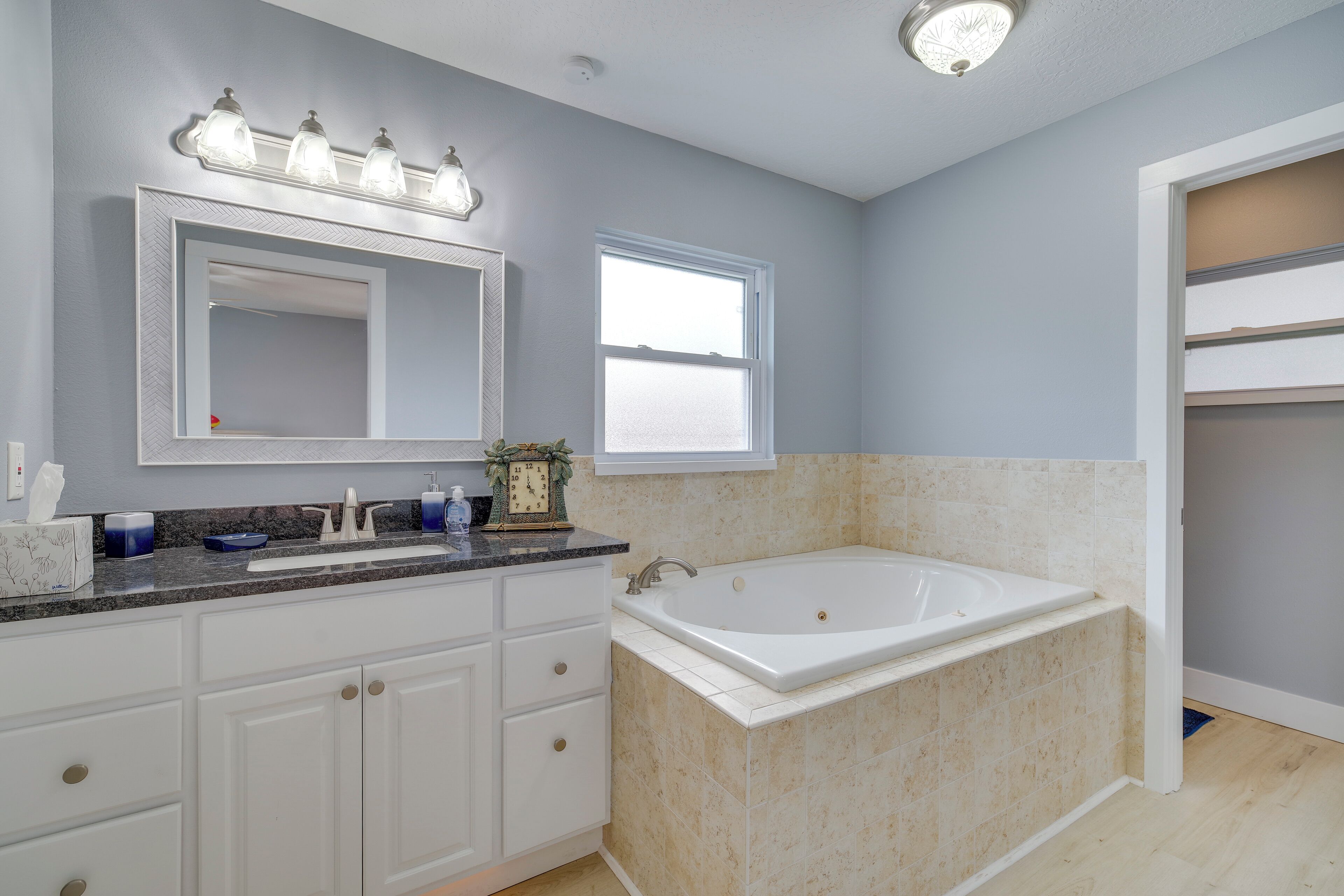 A bathroom with a large bathtub, a vanity with a mirror and lights, and a window.