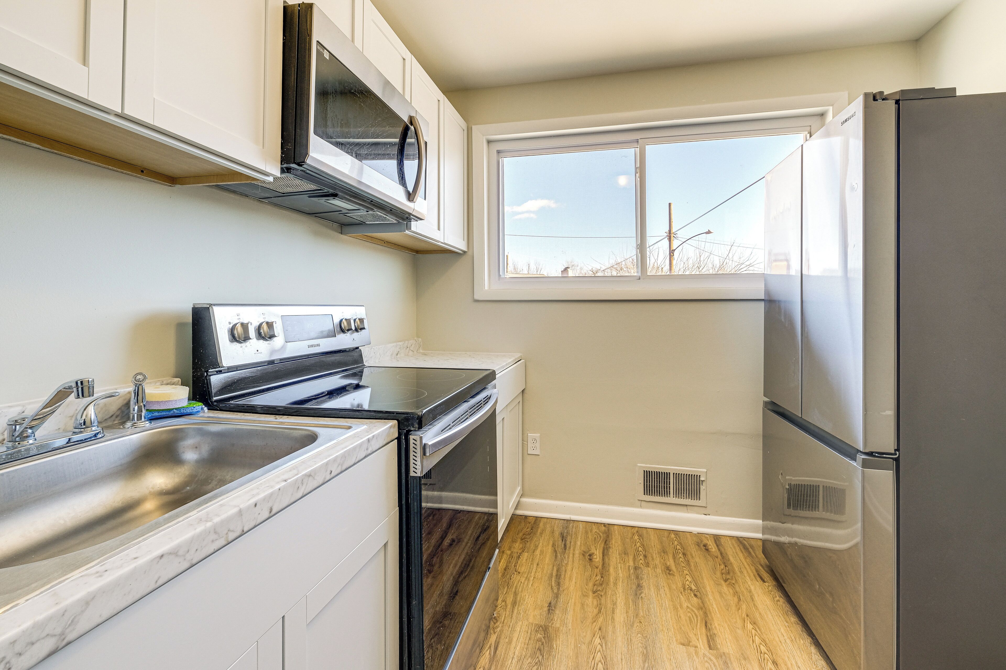 A kitchen with white cabinets, a stainless steel refrigerator, a microwave, and a window with a view of the outside.