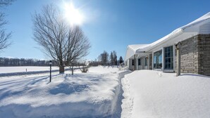A snow-covered landscape with a stone building featuring large windows and a snow-covered roof. - Deck & Fire Pit: Cozy Escape in Morrisville! (Morrisville)
