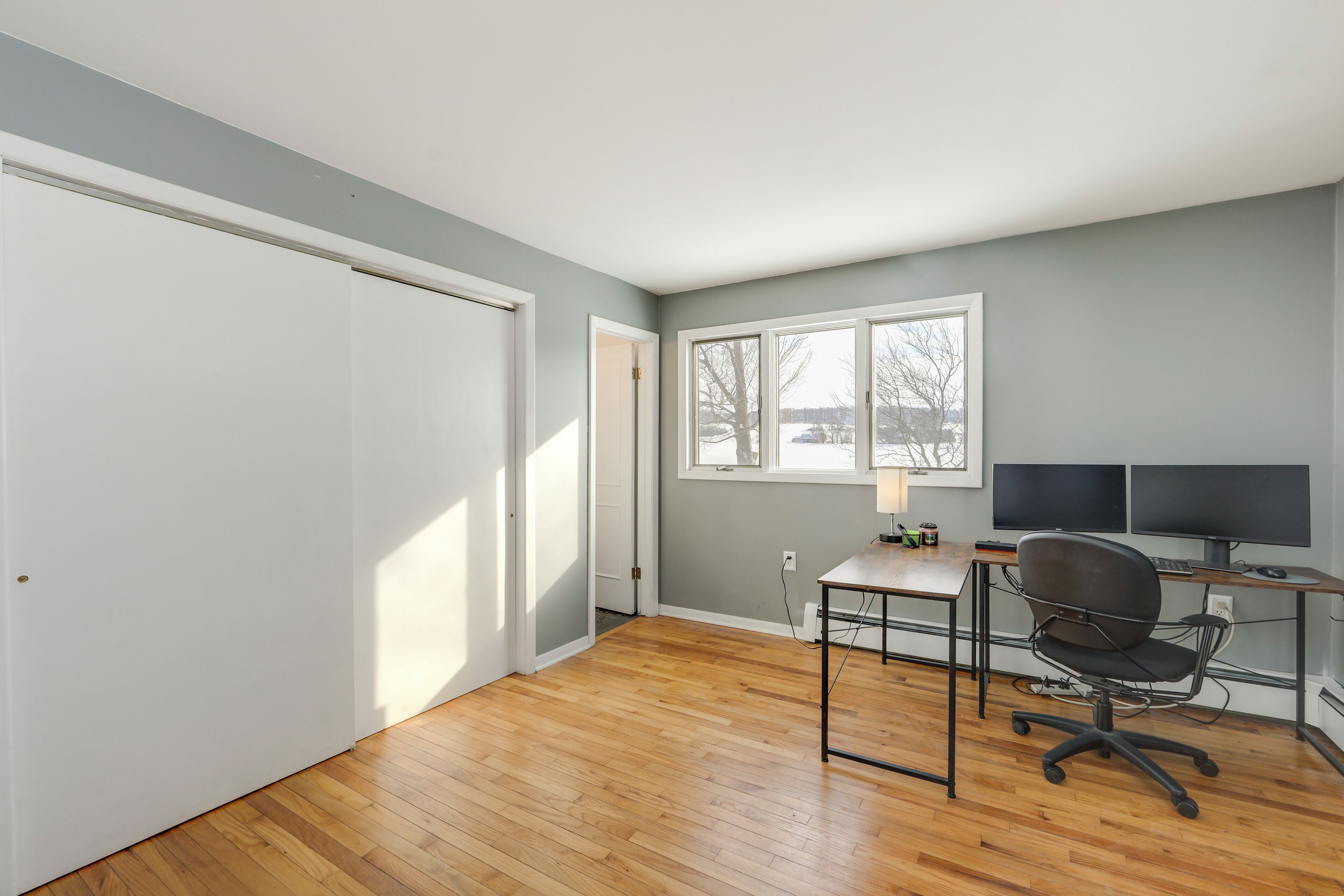 A room with a wooden floor, a desk with a chair, a computer, and a window with a view of trees and a building.