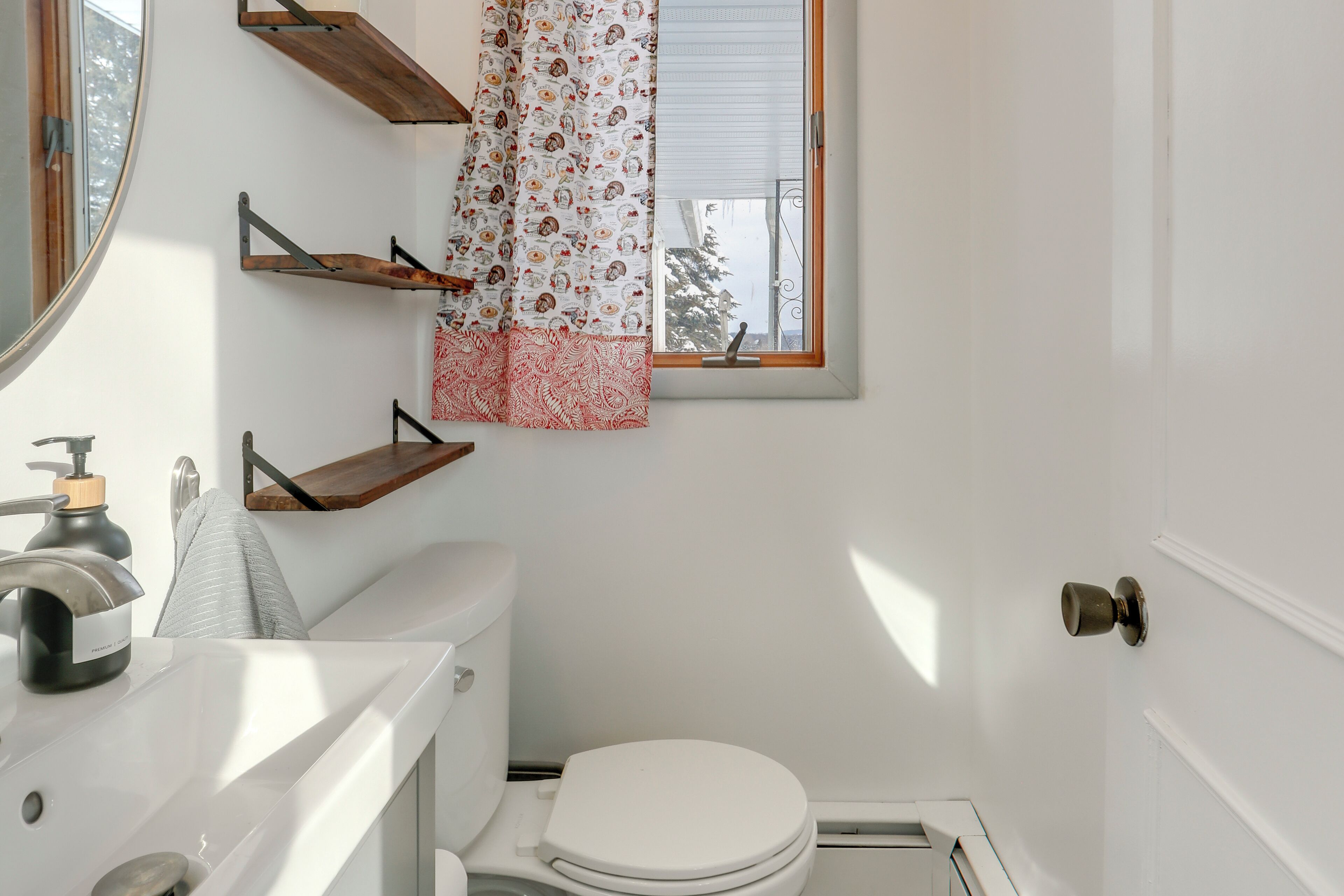 A bathroom with a toilet, a sink, and a window with a curtain. There are wooden shelves above the toilet.