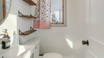 A bathroom with a toilet, a sink, and a window with a curtain. There are wooden shelves above the toilet.