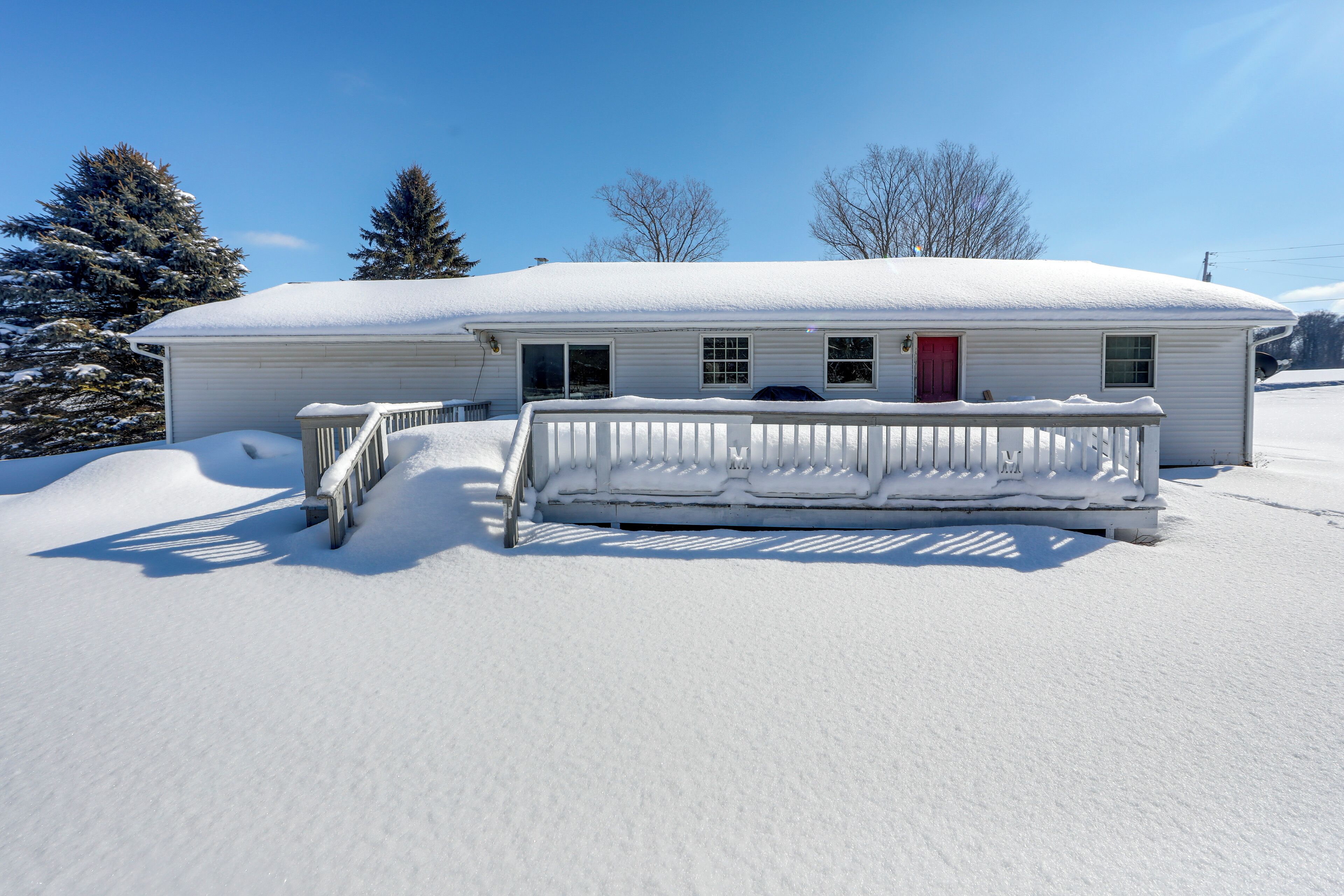 A single-story house with a covered porch and a red door, surrounded by snow.